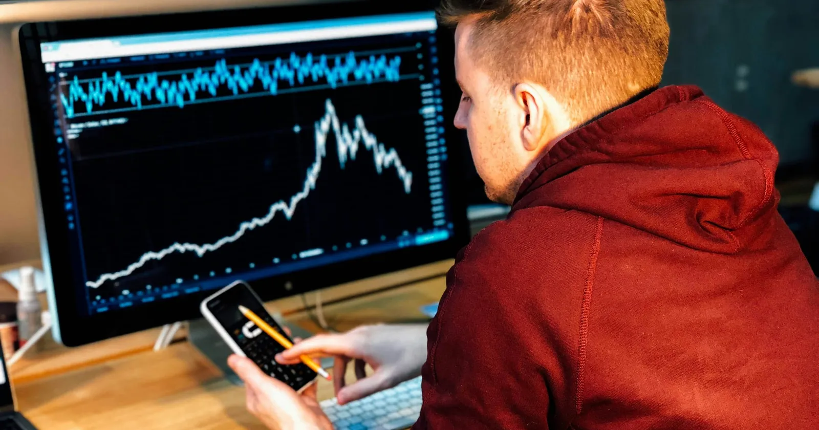 An image of someone on his phone in front of a computer looking at a stock trading application